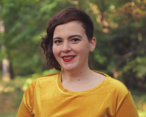 Picture of Annie Tulkin, asymmetrical haircut, gold shirt, smiling, read lipstick.