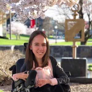 Photo of Dana Jones, young woman with long brown hair, smiling, sitting outside in her wheelchair