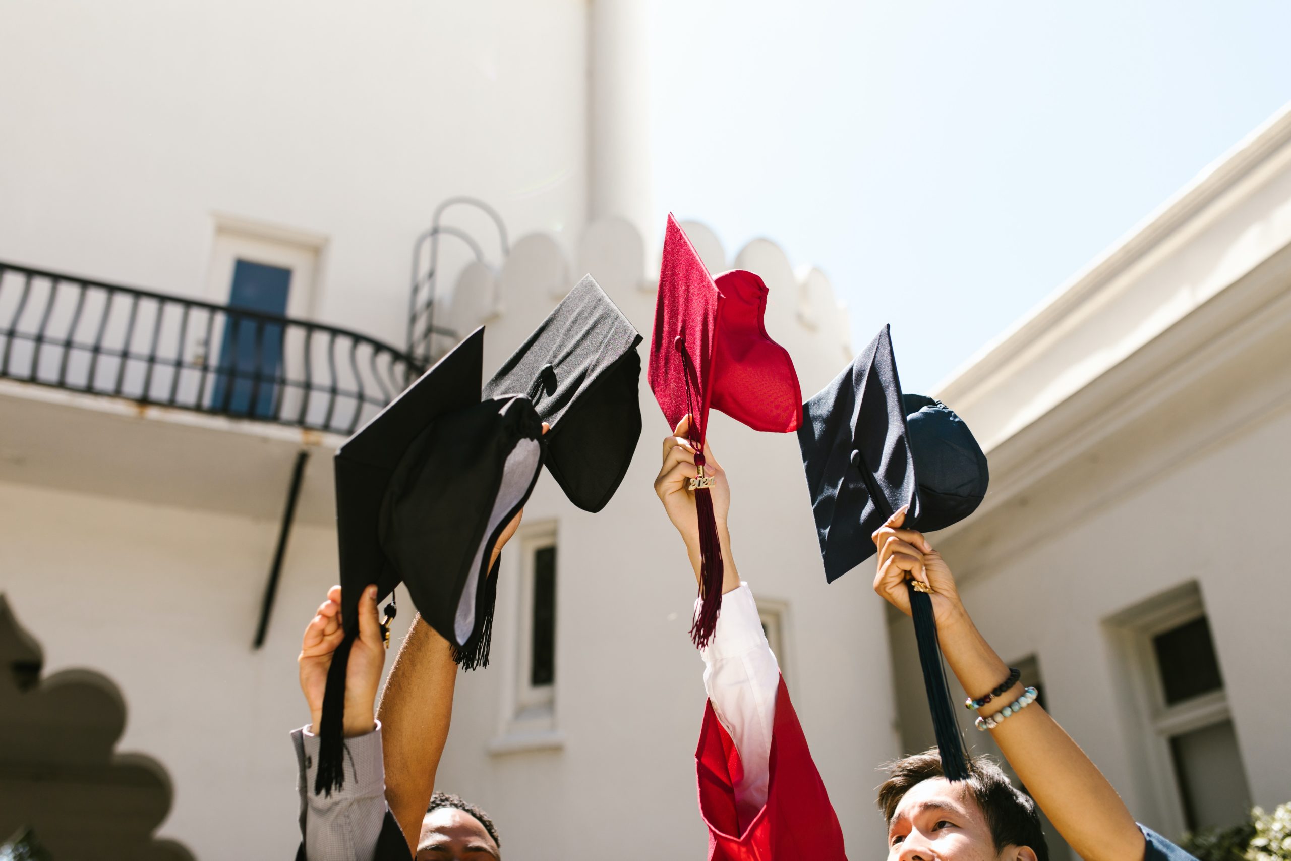 Four arms and hands (brown and white skin tones) holding up graduation caps. One of the caps is red.
